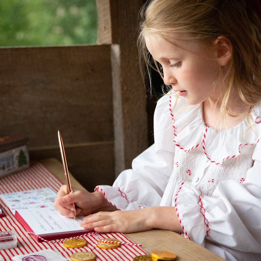 Young girl in a white dress with red trim writing thank you cards at a table with cookies and a striped cloth.