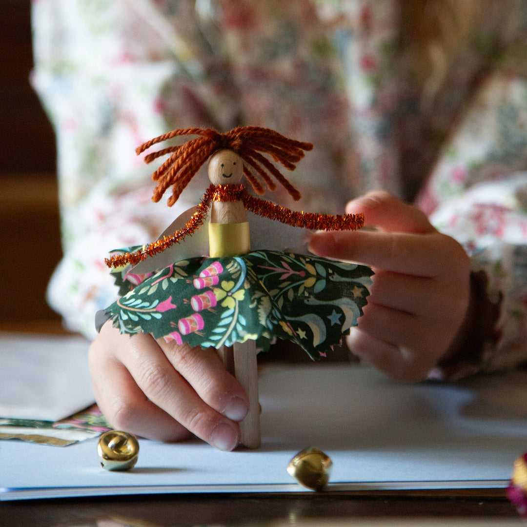 Child holding a handmade wooden doll with a floral dress, surrounded by small objects on a table.