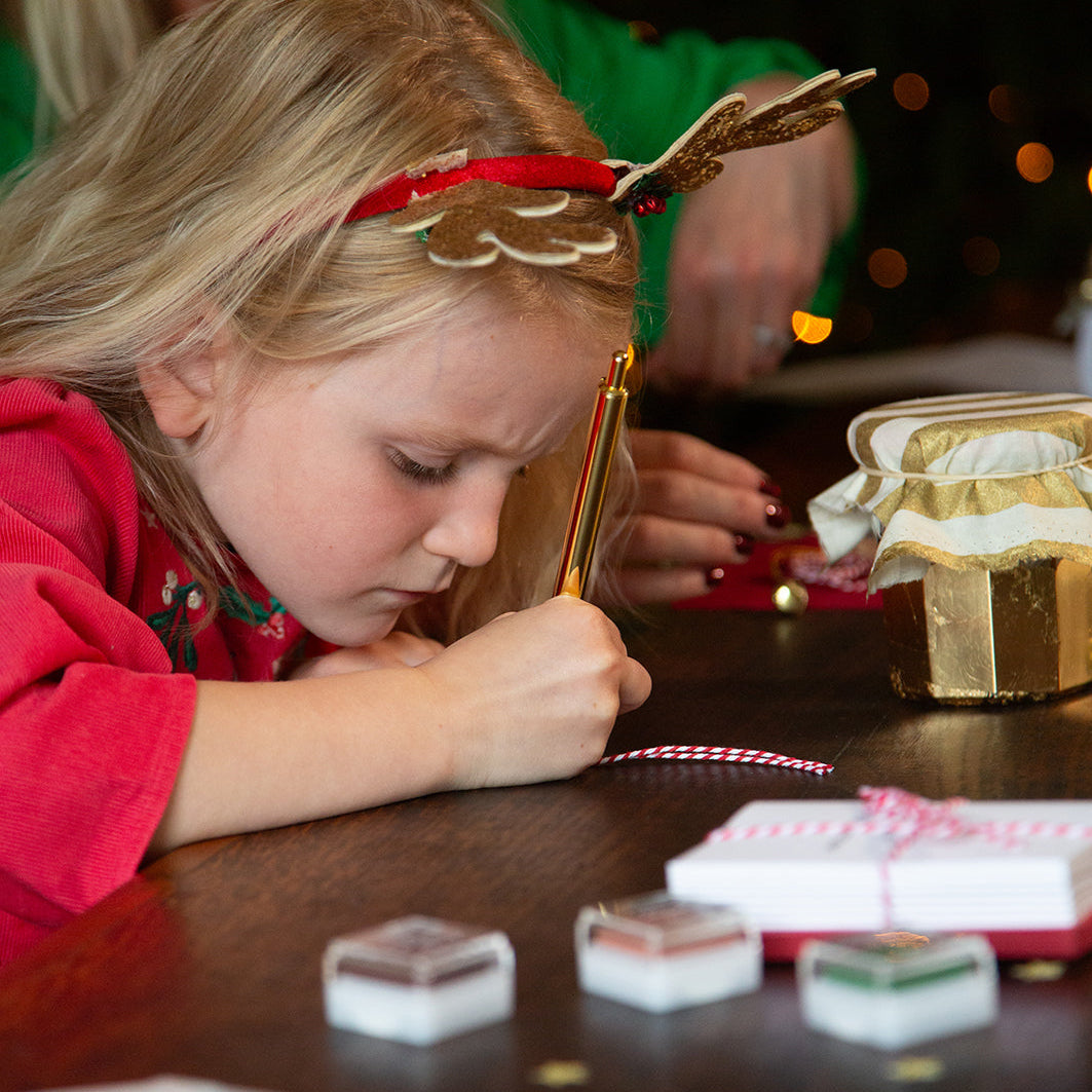 Child writing gift tags at a table with a decorated Christmas tree in the background