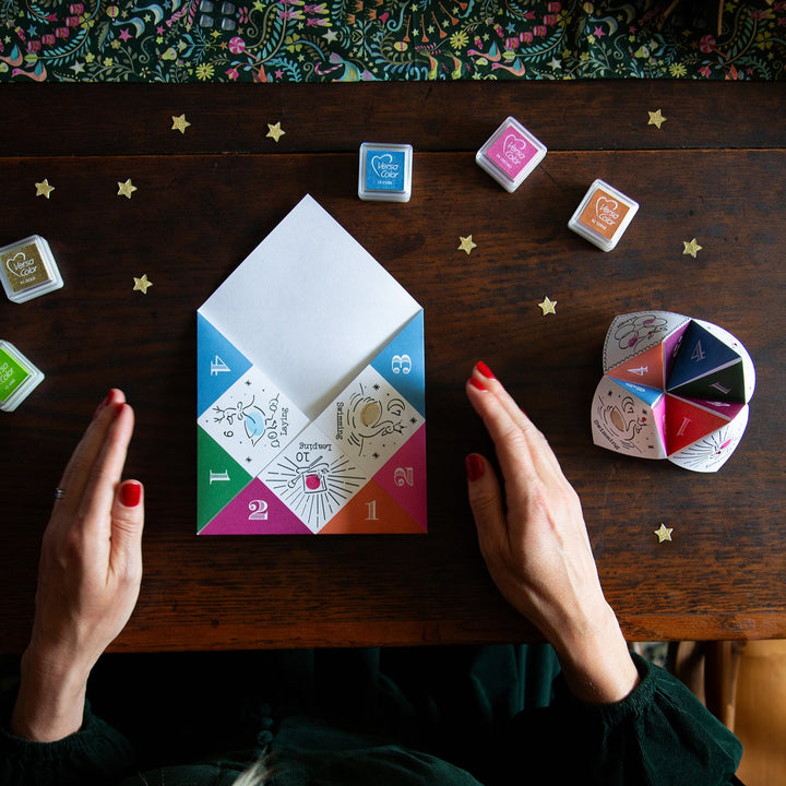 Person holding a colourful Christmas joke teller on a wooden table with various small boxes and a decorative plate.