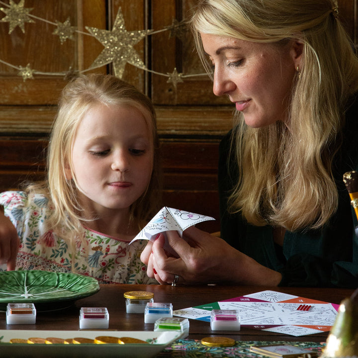 Woman and young girl sitting at a Christmas table with decorative plates and cards, crafting and playing a game.
